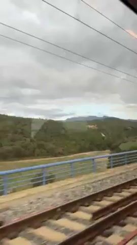The video shows a train journey through a hilly, wooded landscape under a cloudy sky. The camera is inside the train, looking out the window. The initial view includes the train tracks, a blue railing alongside the tracks, and dense green foliage stretching out to the horizon. In the distance, a large, older building with a yellowish roof can be seen nestled within the trees, with a winding dirt road leading toward it. As the train moves, the view shifts to a broader panorama, revealing mountains in the background, still framed by the green landscape in the foreground and more of the building. The final shot zooms in on the mountains, which are slightly obscured by haze. The overall tone of the video is tranquil and observant.