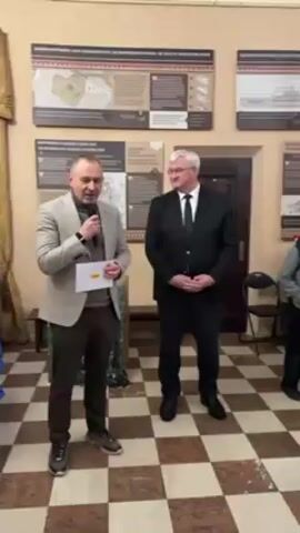 Two men stand in a room with checkered floors and walls covered in informational displays. The man on the left, named Lev Zhydenko, is holding a microphone and a piece of paper, addressing the camera. He is dressed in a gray blazer, dark turtleneck, and dark pants. The man on the right wears a dark suit and tie. They are standing near a doorway and a small table.