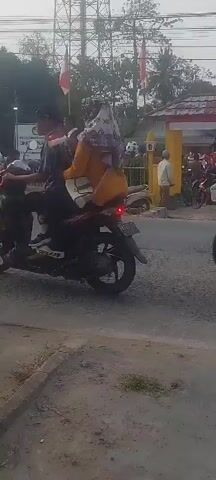 The video captures a street scene with several motorcycles and people. In the foreground, a black motorcycle with a rider is moving to the right. In the background, a group of people is gathered near a building decorated with red and white banners, which appear to be Indonesian flags. Several other motorcycles are parked or moving. A white scooter is visible on the left side of the road. The environment seems to be a public space, possibly a street in a town or village, with trees and utility poles in the background. The overall impression is one of a lively outdoor event.