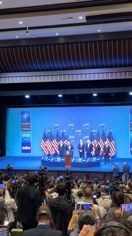 A man, appearing to be Donald Trump, stands at a podium on a stage, speaking to an audience. He is wearing a dark suit and a yellow tie. Behind him, several other men in suits stand in a line. Behind them, American flags are displayed. The backdrop is a blue screen with the NATO and Ankara logos. The stage is set up for a press conference or a formal address, with numerous cameras and microphones in front of the stage, suggesting a media presence. The overall scene depicts a formal political or diplomatic event.