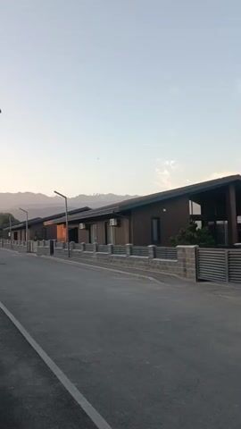 A drone shot slowly pans to the left, revealing a row of modern, single-story houses with dark brown exteriors and sloped roofs. The houses are set back from a paved road, separated by low, grey brick walls and metal fencing. Streetlights with minimalist designs line the road. In the distance, mountains with snow-capped peaks are visible against a pale sky. The scene is peaceful and appears to be a residential neighborhood. The video begins and ends with no discernible action.