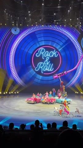 Onstage, a bright Rock & Roll stage show captivates viewers. A large, illuminated blue record with a pink neon Rock & Roll title acts as the backdrop.  Two pastel vintage cars sit center stage, carrying performers in colorful costumes.  To the right, a group of acrobats balances in a human tower on top of a bicycle. Adjacent to them, smaller bicycles, one carrying a child with a dog passenger, and the other pulling a dog on a leash, add to the lively spectacle. The audience, visible at the bottom, is enjoying the performance.  Bright yellow beams of stage lighting illuminate the scene.