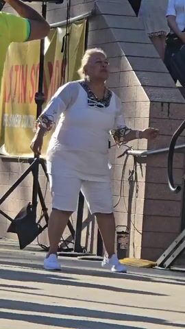 A woman in a white outfit dances outdoors. She wears a white top with sheer sleeves, white shorts, and white sneakers. She has a colorful necklace and earrings. Behind her, a yellow banner with text is visible, as well as a speaker on a stand. A person in a yellow shirt is to her left, and another person in a white shirt is to her right, further up some stairs. The ground is paved and sunlit, with long shadows.