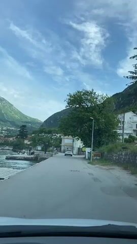 The camera is mounted in a car driving on a narrow road alongside a bay. The bay is bordered by a stone wall and has mountains rising sharply in the background. Several boats, including a sailboat, are in the water.  The sky is mostly clear with wispy clouds. On the right side of the road is a building and trees. As the car drives, it passes a group of three people sitting on the grass near the water's edge. One person is using a camera mounted on a tripod.
