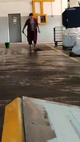 The video shows a person mopping a floor in what appears to be a warehouse or industrial setting. The person is wearing a maroon robe-like garment, dark pants, and blue gloves. They are holding a mop and moving it back and forth across the floor. To the left of the person is a green bucket, and to the right, there is a metal cart with white bags on it. In the background, there is a yellow structure with a red cabinet, a grey door with a green sign, and a blue dumpster. The floor is wet and reflective, indicating it has been recently cleaned. The overall scene is one of routine cleaning in a non-residential environment.