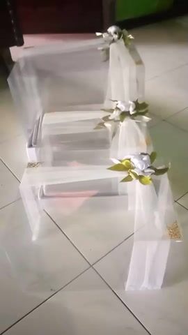 The video shows a close-up of a decorated, multi-tiered object that resembles a wedding cake stand or a display shelf. The object is covered in white tulle fabric, which is draped and tied with white ribbon. Each tier is adorned with white artificial flowers and green leaves. Gold corner protectors are visible on some edges of the tiers. The object is placed on a tiled floor. The lighting is dim, and the image is slightly blurry, suggesting movement or a low-quality camera. The background is out of focus, showing hints of furniture and a green wall.