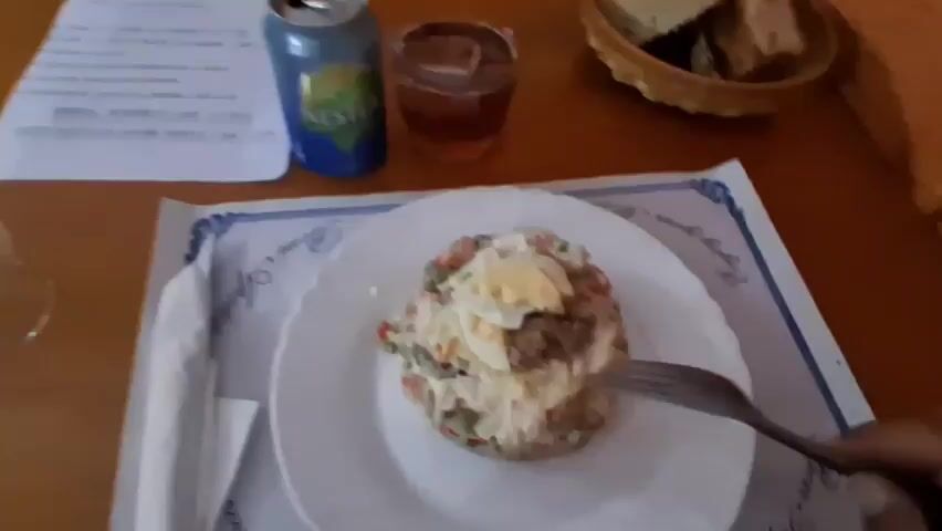 This media file captures a close-up view of a meal being served at a table. The primary subject is a white plate holding a salad with visible ingredients like chopped vegetables, what appears to be tuna, and hard-boiled egg. A fork is actively digging into the salad, suggesting the meal is in progress.

To the left of the plate, a can of "Nestea" beverage is visible, along with a glass of dark liquid, likely tea or iced coffee. A folded white napkin sits to the left of the plate. In the background, a basket contains slices of bread. A menu or document with text is also on the table, partially visible, with bullet points and some Spanish text, including "MENU: MENU CANASTRO POSTRE Y CAFE 12.00 €".

Further in the background, the scene shifts to a more interior view of what appears to be a restaurant. An orange wall is adorned with a vibrant, colorful painting of a person with a headdress. The lighting suggests an indoor setting, possibly during the day or evening, with warm tones. There are hints of other tables and patrons in the blurred background, indicating a public dining establishment. The overall impression is a casual meal in a Spanish setting, with emphasis on the food and beverage.