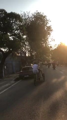 The video captures a street scene at sunset. The foreground shows the side of a small food cart with its wheels visible. A person in white is riding a tricycle-like vehicle with a canopy in the middle of the road, heading away from the camera. To the left of the road, a dark-colored sedan is parked. Beyond the parked car, a fence and some buildings are visible. The road itself is paved, and there are several motorcycles riding in the distance. Tall trees line the right side of the road, and the setting sun casts a warm glow across the scene. The overall impression is one of a busy street in an urban or semi-urban environment during the late afternoon or early evening.