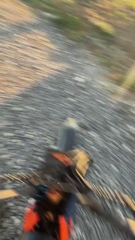 Here is a brief description of the video:

The video shows a person wearing a black helmet and a blue and black long-sleeved shirt standing on a gravel road. The camera is attached to the person, providing a first-person perspective. The background includes a building, grass, and a clear blue sky. The camera moves around, showing different angles of the surroundings.
