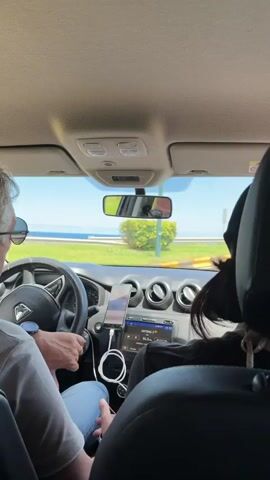 The video shows a view from the back seat of a car, focused on the driver. The driver is an older man with graying hair, wearing sunglasses, a light-colored shirt, blue jeans, and a watch on his left wrist. He also has a hearing aid in his left ear. He's driving on a sunny day, with a clear blue sky visible through the windshield. The car's dashboard is visible, featuring a smartphone mounted in a holder, connected with a charging cable. The car's radio displays "ANTENA 3" and "91.3 POP". Outside the car, palm trees and a glimpse of the sea can be seen through the windshield. Another person is partially visible in the back seat, to the right of the frame.