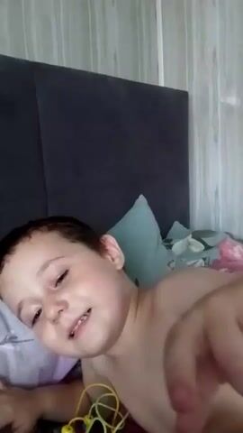 A close-up video shows a young boy lying in a bed with a grey headboard behind him. The boy is looking at the camera with a slight smile. He has short, dark hair and appears to be wearing a shirt. In the foreground, a child's toy with colorful beads and wire is visible. The room appears to be a bedroom, with a patterned pillow and a window with sheer curtains in the background. The lighting is bright, suggesting it is daytime. The camera appears to be held at a slight downward angle, capturing the boy from his chest up.