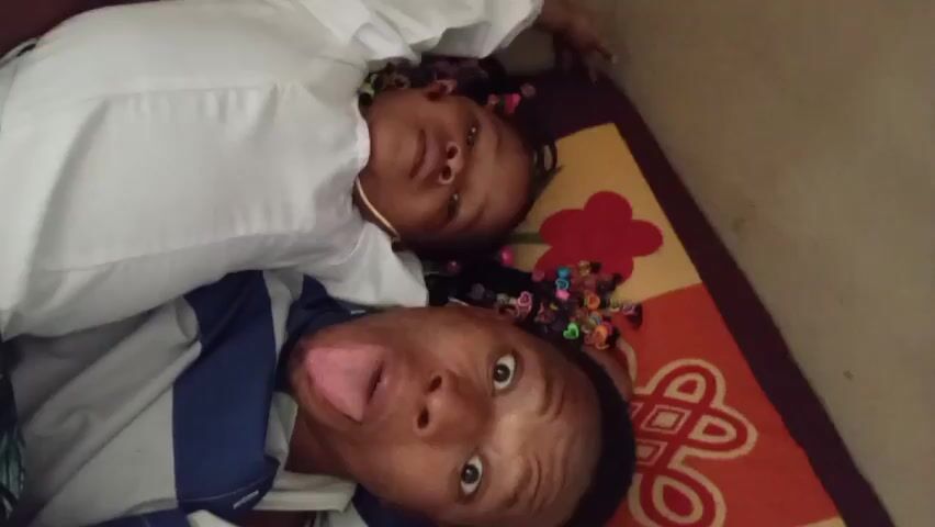 A person with short, dark hair and a white and blue striped shirt is lying down next to a young child wearing a white shirt. The child has dark hair with colorful beads and a gold necklace. The person lying next to the child sticks their tongue out and opens their mouth wide. The child also sticks their tongue out. They are lying on a multi-colored rug with floral designs. The background is a plain wall.