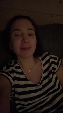 A woman is lying down and smiling at the camera. She is wearing a black and white striped t-shirt and a necklace. She has short, dark hair. The background is dark and blurry. She moves her head and touches her neck.