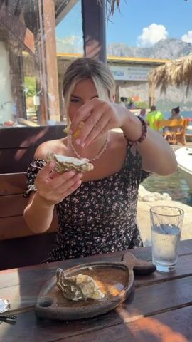 A woman with blonde hair pulled back, wearing a black floral print dress and a pink beaded necklace, sits at a wooden table outdoors. She is in the process of eating an oyster, squeezing a piece of lemon juice over it with her right hand. Her left hand holds the oyster. In front of her on the table is a small wooden board shaped like a fish, with another oyster on it. To her right, a glass of water sits on the table. Behind her, a body of water is visible, with mountains and buildings in the distance. Sunlight illuminates the scene.