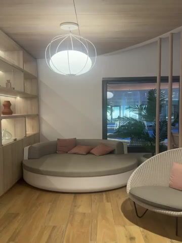 The video shows a corner of a room with a round daybed, a potted plant, a chair, and a large window overlooking an indoor pool. The daybed is light beige with three rose-colored square pillows. A white orb-shaped light fixture hangs above it. Next to the daybed is a white wicker chair with a light gray cushion. A tall potted palm stands between the daybed and the window. The window offers a view of a large indoor swimming pool with people swimming and walking around. The walls are white, and the floor is wooden. There is also a built-in wooden shelving unit on one side of the room. A wood slat divider partially frames the shot.