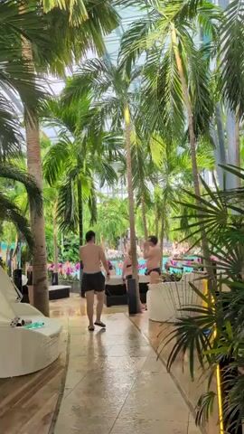 This is a first-person perspective video shot in a tropical-themed indoor resort or water park. The camera is positioned at eye level and is moving forward along a tiled walkway. The walkway is lined with lush green palm trees of varying heights, some with large fronds extending over the path. To the left, there are white lounge chairs and what appears to be a white sectional sofa. To the right, there are more palm fronds and some modern-looking white seating. In the background, visible through the trees, is a large pool area with many people swimming and lounging. Several individuals are walking away from the camera down the path, also heading towards the pool area. One person in the foreground is wearing black swim trunks and has long dark hair. They are carrying a dark blue piece of clothing in their left hand. Another person further down the path is wearing a black bikini. The overall atmosphere appears to be relaxed and recreational. The ceiling of the structure is not visible, but the lighting suggests it is either daytime or well-lit artificial lighting.