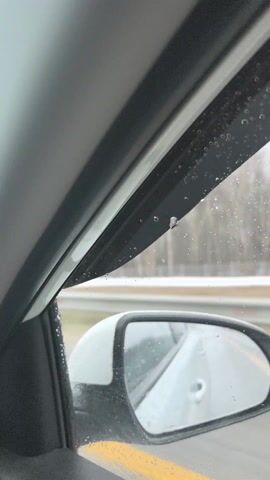 The video shows the view from inside a car driving on a road, with rain on the car window and side mirror. There's a line of trees visible in the distance. Music is playing in the background. The focus is on the rain droplets on the window and the passing scenery.