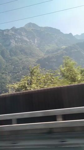 The video shows a view from a moving vehicle. In the foreground, there is a black barrier with a silver railing on top. The barrier is wide and appears to be separating the vehicle from a lower level. Above the barrier, there are trees and greenery that are blurred due to the motion of the vehicle. In the background, a large, imposing mountain rises under a clear blue sky. The mountain is covered in dense green vegetation on its lower slopes and rocky, lighter gray surfaces higher up. Several power lines are visible crisscrossing the sky in front of the mountain. The lighting suggests it is daytime, likely morning or afternoon, with the sun casting a bright light on the scene.