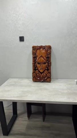 A video shows a carved wooden mask on a table. The mask is rectangular and features a stylized face with elaborate scrollwork and floral motifs around it. The mask is brown and appears to be varnished. The table is light-colored and has a textured surface. In the background, there is a wall with a textured finish and a light switch. The lighting is even and appears to be from an artificial source. The camera moves slightly, zooming in on the mask. There are no people visible in the video.
