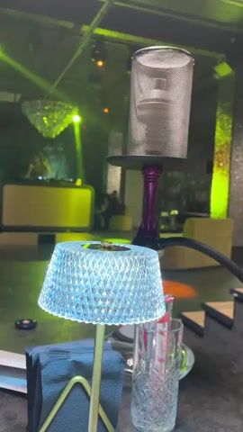 The video showcases a hookah in a dimly lit, possibly indoor, entertainment venue. The hookah itself is the primary subject, a modern design with a purple stem and a metallic mesh cover over the bowl. Smoke is actively being produced and rising from the hookah. In the foreground, a small, crystal-like decorative lamp with a textured surface is placed on a table, alongside a napkin holder and a glass.

The background reveals a vibrant scene typical of a club or lounge. Green and yellow lights are projected, creating an atmospheric ambiance. A DJ booth with a yellow illuminated surface is visible, suggesting music is being played and a DJ is present. In the distance, other patrons can be seen, though they are out of focus. The overall impression is a social gathering at night, likely in a city like Magnitogorsk, Russia, where people are enjoying a night out with music and hookah. The lighting and the presence of a DJ imply a lively and possibly energetic atmosphere. There is no visible text in the video.