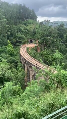 The video shows an aerial view of a curved railway bridge made of stone arches. The bridge is surrounded by lush green forest and trees. People are walking on the railway tracks of the bridge. In the background, a tunnel is visible in a hillside. The sky is overcast with clouds.