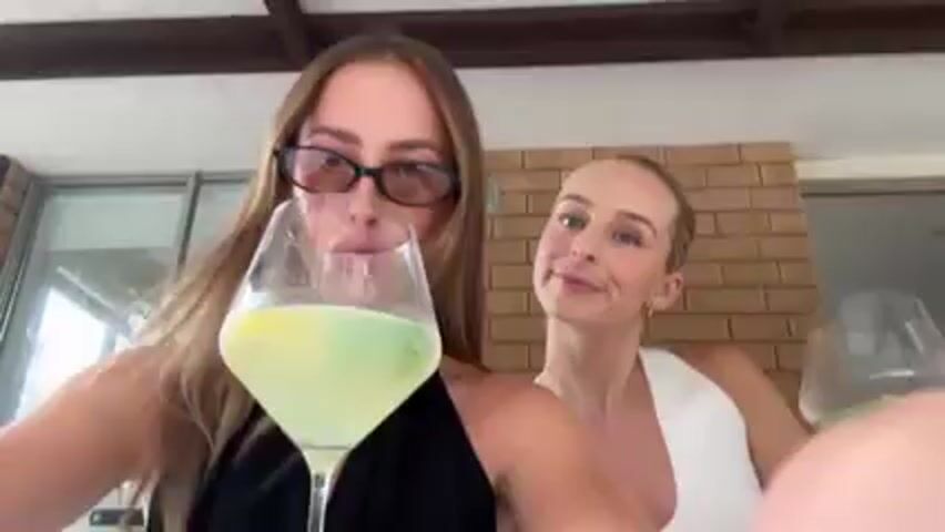 Two young women are standing close together, holding stemmed glasses with a pale green liquid. The woman in the foreground has long brown hair, glasses, and a nose ring, and is wearing a black top. The woman in the background has blonde hair pulled back, and is wearing a white top. Both are smiling slightly. They are outdoors on a patio, with a brick wall behind them and a window to the left. Music is playing in the background.