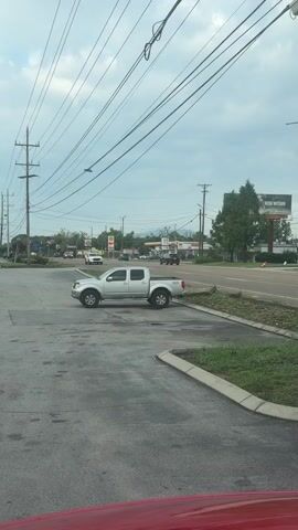 A silver pickup truck is parked in a parking lot with its hood up. In the background, there is a street with traffic, gas stations, and a billboard. The sky is cloudy. A red object, possibly the hood of another vehicle, is visible in the foreground.