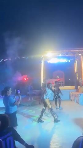 The video shows an outdoor nighttime event with a stage in the background. A band or performers are on the stage, illuminated by stage lights and what appears to be fog or smoke. In the foreground, several people are present. One person is taking a photo with a phone. Another person is dancing or moving in a lively manner. Two balloons are visible in the sky. Fireworks are also seen exploding in the sky. The atmosphere appears festive.