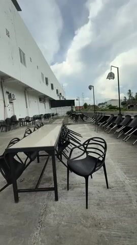 The video pans across an outdoor seating area. In the foreground, a long, narrow table with a light-colored surface is set up with black chairs. Several chairs are placed around the table, and some are stacked. Further back, a row of black chairs is neatly arranged along the edge of the seating area, with their backs facing the viewer. Beyond the chairs, there is a building with a white exterior and several windows. In the distance, there are trees, power lines, and a cloudy sky. The overall scene suggests an empty outdoor dining or gathering space.