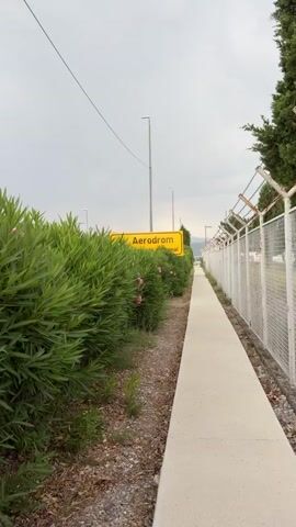 The media file shows a yellow sign with black text that reads "Aerodrom" and an arrow pointing left. Below "Aerodrom," it says "meuna'rional terminal." The sign is placed next to a lush green hedge with some pink flowers. To the right of the hedge is a concrete walkway. Beyond the walkway, there is a white metal fence with barbed wire on top, marking a perimeter. The scene appears to be at an airport, with tall light poles visible in the background. The sky is overcast, suggesting cloudy weather. The time of day appears to be daytime, possibly late afternoon or early morning due to the diffused light. The overall mood is neutral, focused on the signage and the airport environment. The location context is Golubovci, Montenegro.