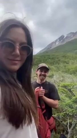 A video shows two people, a woman in the foreground and a man behind her, amidst lush green vegetation. The woman wears sunglasses and a white sweater. The man wears a baseball cap and a dark T-shirt, holding a red backpack. The camera pans around them, revealing a mountainous landscape with a snow-capped peak in the background. The weather appears overcast. The people appear to be on a hike.