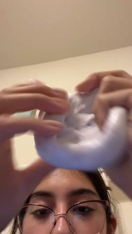 The video shows a person with brown hair and glasses holding and stretching a white, shimmery substance, presumably slime, above their head. The background includes a light beige wall, a white headboard of a bed, and a small framed painting. The person's hands are visible as they manipulate the slime, stretching it into a thin, translucent sheet. The camera remains stationary, capturing the scene from a low angle, looking up at the person and the slime.