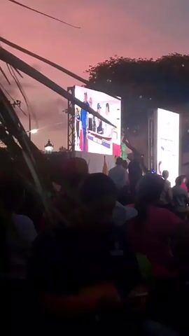The video shows a gathering of people at an outdoor event at dusk. Two large screens are visible, one showing a video. The crowd is silhouetted against the pink and purple sky, and lampposts are interspersed among them. People are standing and moving around. Palm fronds frame the shot on the left. The event appears to be taking place in a public space.