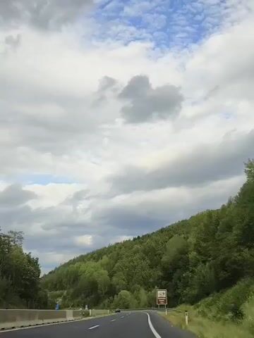 The video shows a roadside view from a moving vehicle. The camera is pointed forward, capturing a highway stretching into the distance. On the right side of the road, a sign with an image of a castle and text, including "Hochschloss Gloggnitz 1300m," is visible. The landscape is dominated by lush green trees covering a hillside that slopes upwards to the right. The sky above is partly cloudy with a mix of white and gray clouds. The road has white lane markings and a concrete barrier on the left.