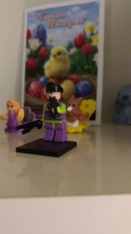 The video shows a group of small figurines displayed on a shiny, light-colored surface. There is an Easter card in the background featuring a yellow chick, tulips, and colored eggs. The figurines include a Lego figure holding a gun, a snowman on a snowboard, a reindeer, a Green Lantern figure, and a Rapunzel figure. The camera moves slightly to provide a better view of all the figurines. The lighting in the scene is soft and diffused.