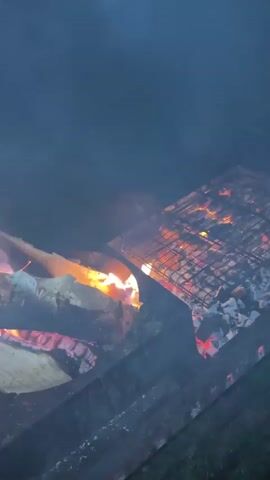 The video shows a charcoal grill with glowing embers and burning logs. A metal grate is positioned over the coals, with what appears to be food on skewers being cooked. Smoke rises from the grill, obscuring the background. The setting appears to be outdoors at night, with trees and vegetation visible in the dim light. In the background, there is a structure with lights and a hint of fencing.