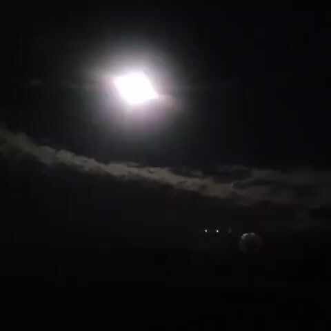 The video begins with a bright, rectangular light source in a dark environment. The light pulsates and flares, creating lens flares. A dark, textured surface is visible below the light. The camera pans slightly, causing the light to stretch and distort. In the lower right corner of the frame, a small, metallic object, possibly a can, is visible. The scene is dark and appears to be outdoors at night. The lighting is intense and artificial.