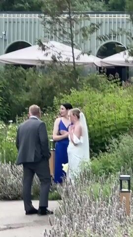 A man in a suit stands facing a woman in a white wedding dress. Another woman in a blue dress stands behind the bride. The scene appears to be outdoors in a garden setting with lush greenery and flowering plants. A building with white awnings is visible in the background. The people are standing still and appear to be in conversation.