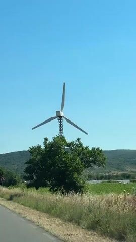 The video shows a wind turbine with three blades against a clear blue sky. The turbine is situated behind a tree and in front of a green, hilly landscape. A road with a grassy verge is visible in the foreground, and a triangular warning sign with an arrow indicating a curve is present on the side of the road. The turbine blades are slowly rotating. The video is approximately 4 seconds long and appears to be filmed from a moving vehicle.