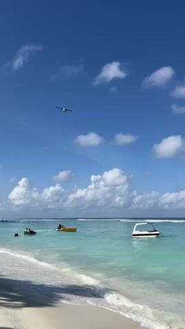 A seaplane flies across a bright blue sky with scattered white clouds. The plane is white and blue with two propellers on its wings. Below the plane, a turquoise ocean meets a sandy beach. Several small boats are in the water, one of which is white and modern, while others appear to be smaller dinghies. Two people are visible in the water near one of the dinghies. The overall scene is serene and tropical.