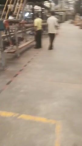 The video shows two men standing in what appears to be a factory or industrial setting. The floor is concrete, and there are red and black lines marking a boundary on the ground. In the background, there are various machines and equipment, including stairs leading up to a higher level. The man on the left is wearing a yellow shirt and dark pants, and he is facing away from the camera, looking at some equipment. The man on the right is wearing a white shirt and brown pants, and he is also facing away from the camera, walking slowly to the right. The lighting in the video is somewhat dim, and the overall atmosphere seems to be that of a working environment.
