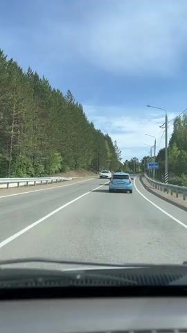The video shows a view from inside a vehicle driving on a paved road. The road has white lane markings and is surrounded by a dense forest of tall, green trees on the left side. On the right side of the road, there are guardrails and more trees. The sky is blue with scattered white clouds. Two cars are visible on the road ahead. A white car is in the left lane, and a blue car is in the right lane. As the vehicle in the video moves forward, the blue car in front of it also moves forward. The white car in the left lane appears to be stationary or moving much slower. The blue car is further ahead in the right lane. Another car, possibly red, is now visible behind the blue car. The perspective is looking forward through the windshield.