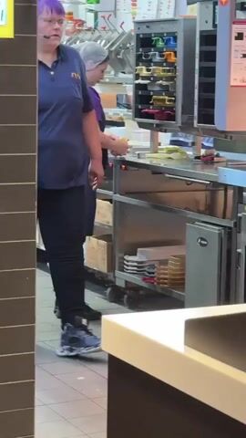 The video shows a fast-food kitchen environment. In the foreground, a person wearing a blue polo shirt and black pants is walking away from the camera towards the right. Their face is not visible. In the background, another person wearing a purple shirt with a logo, black pants, and a white hairnet is working at a stainless steel counter. They are reaching for something on a shelf. The environment includes stainless steel equipment, shelves stacked with containers, and a tiled floor. The lighting appears to be fluorescent and typical of a commercial kitchen.