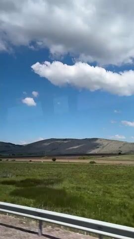 The video shows a daytime landscape viewed through a vehicle window. A field of green grass stretches towards a brown flatland, behind which rise low, grey hills. The sky is bright blue, dotted with white, puffy clouds. A grey guardrail runs along the bottom of the frame, indicating that the vehicle is on a road. The image appears to be captured on a bright, sunny day.