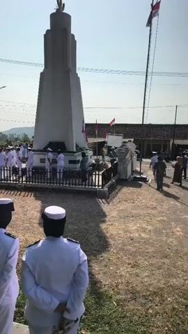 The video captures an outdoor ceremony at a monument. Several people in white uniforms stand at attention in front of and around the monument, which has a unique, tiered design topped with a cactus-like sculpture. Some individuals are gathered in groups, and a flag pole with a flag can be seen in the background. The ground is dirt and gravel, with some vegetation and trees present. The sky is clear and blue, suggesting it is daytime. Three individuals in white uniforms are in the foreground, facing away from the camera, towards the monument.