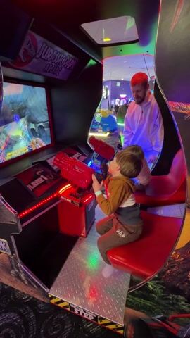 A father and two young boys are playing a Jurassic Park arcade game in a brightly lit arcade. The boy in the foreground is seated and holding the red gun-shaped controller, actively engaged in the game displayed on the screen. The game shows a car exploding and scenes of dinosaurs and landscapes. The arcade game is bordered by red lights and "Jurassic Park" branding. Behind the boy in the foreground, another younger boy is also seated, but not as actively engaged. The father stands behind the boys, watching them play. Other arcade games and people can be seen in the background. The arcade has colorful lighting and a playful atmosphere. The arcade game's base is a silver, diamond-plated material.