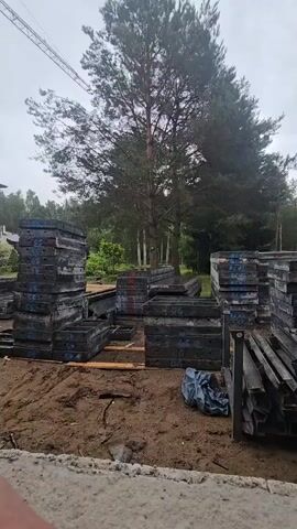 A construction site is shown with stacks of concrete formwork. The formwork is made of dark metal and is stacked in several piles. Some pieces of wood are on the ground between the stacks. A blue tarp is also visible on the ground. In the background, there are trees and a crane. The sky is overcast.