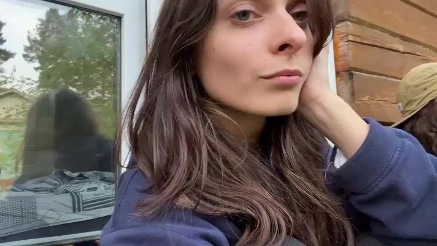 A young woman with long, dark, wavy hair and green eyes is looking at the camera. She is wearing a dark blue hoodie and her left hand is resting on her face. She has a neutral expression. The background appears to be outdoors, with a window reflecting trees and a building. There is also a glimpse of another person wearing a baseball cap and a dark hoodie on the right side of the frame. The woman briefly waves her right hand towards the camera at 00:02.