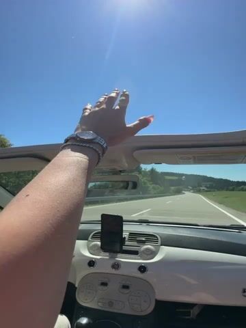A first-person perspective shot shows a person's arm and hand raised toward the sky. The hand is open with fingers spread. The person is in the passenger seat of a white convertible. The car is moving on a road. The sun is shining brightly in a clear blue sky. The interior of the car is visible, including the dashboard and a phone mounted to it. Trees and hills are visible in the background.