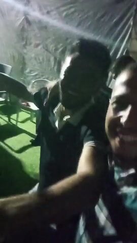 This is a shaky, blurry video of two men. The man on the right is closer to the camera and is smiling. He is wearing a plaid shirt. The man on the left is behind him and appears to be wearing a black collared shirt with white trim. They seem to be outdoors at night, possibly at an event or party, as there are blurred lights and a tarp-like structure in the background. The overall impression is of a candid, informal moment.